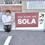 潮江中学校分教室SOLAの看板前で笑顔の中島匠一代表